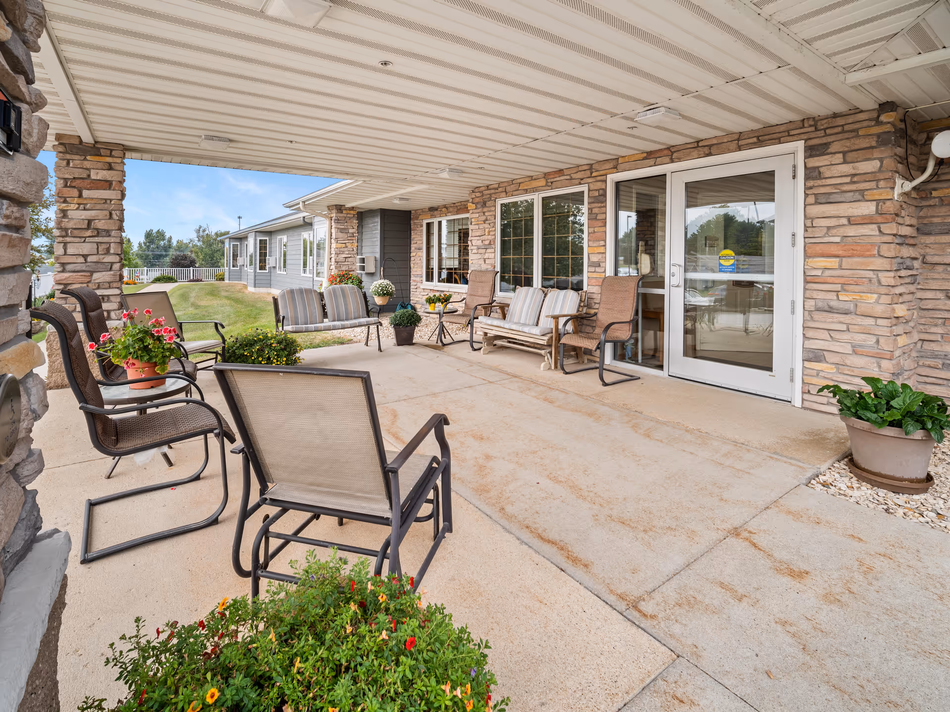 Covered outdoor patio area with several chairs and benches arranged around small tables. The patio is adjacent to a building with stone and siding exterior walls and large windows. There are potted plants and flowers placed around the seating area, and a grassy lawn with trees is visible in the background under a blue sky.