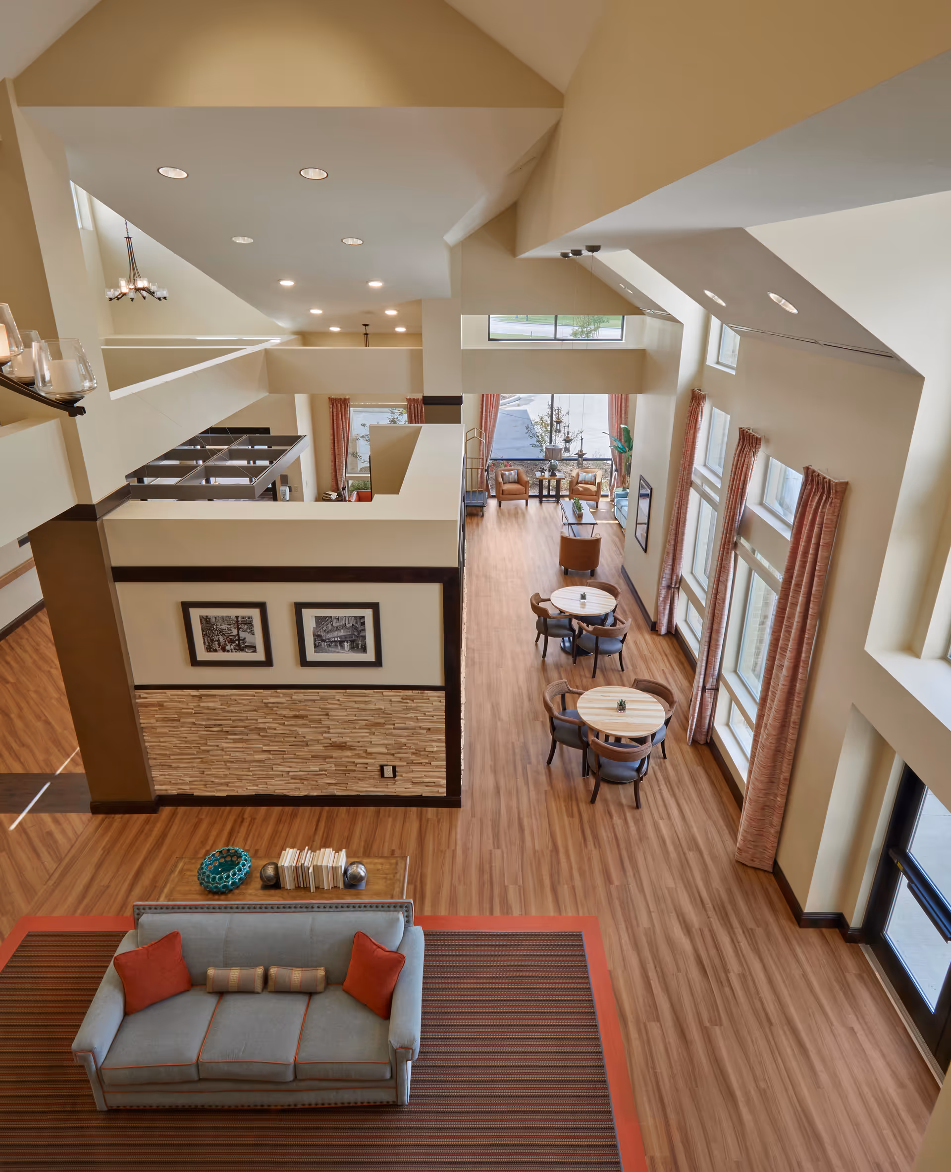 Spacious and well-lit interior of a senior living facility with high ceilings and large windows. The room features a seating area with a gray sofa and red pillows on a striped rug, several round tables with chairs, and additional seating near the windows. The floor is wood, and the walls are painted beige with framed pictures and decorative stone accents.