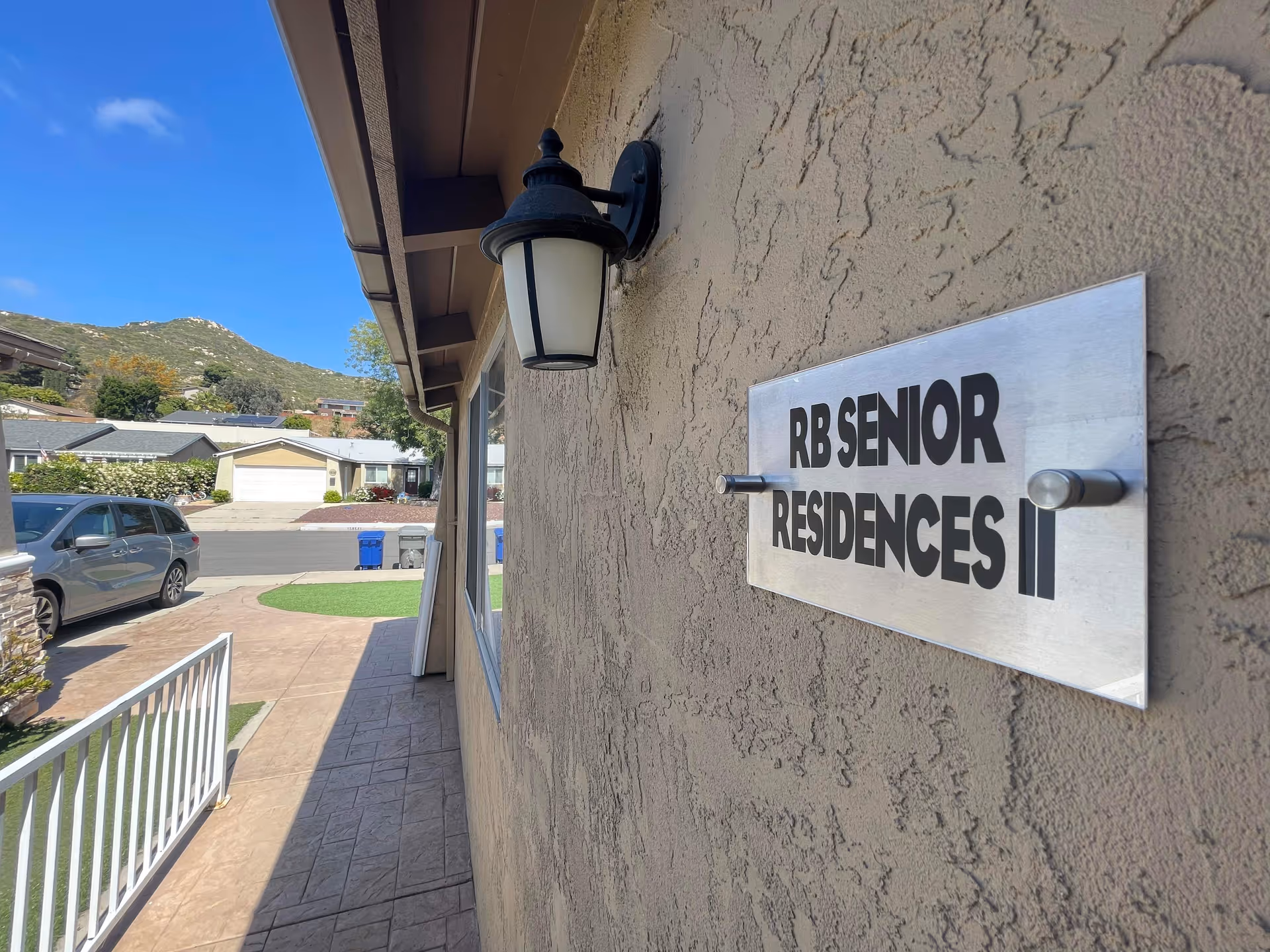 Close-up view of a wall-mounted sign reading 'RB SENIOR RESIDENCES II' on the exterior of a building, with a black outdoor light fixture above it. A driveway, parked car, and neighboring houses with hills in the background are visible under a clear blue sky.
