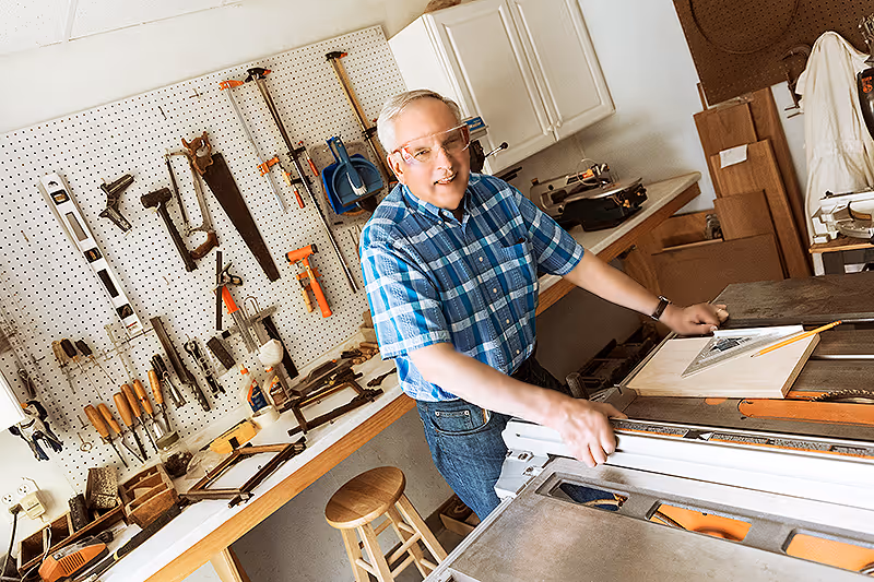 An elderly man wearing safety glasses and a blue plaid shirt is working in a woodworking shop. He is standing next to a table saw with a pencil and a triangular ruler on a piece of wood. The background shows a pegboard with various woodworking tools hanging on it and a wooden stool nearby.