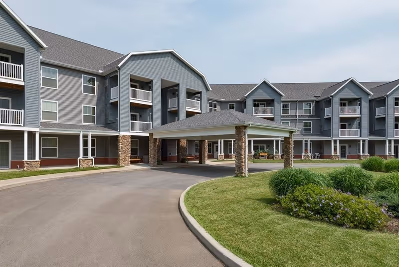Exterior view of a senior living facility with a large covered entrance supported by stone pillars, surrounded by a paved driveway and well-maintained green landscaping with bushes and grass. The building has multiple floors with balconies and windows.