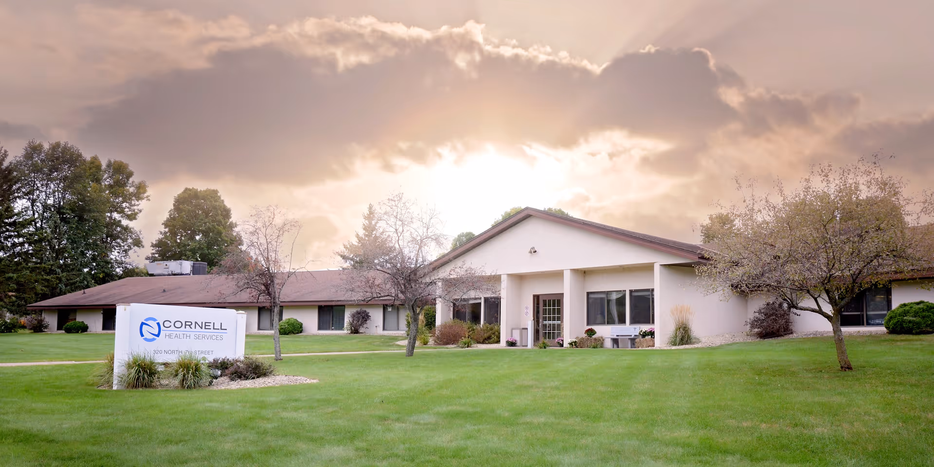 Exterior view of Cornell Health Services building with a large green lawn and trees in the foreground under a cloudy sky with sunlight breaking through.