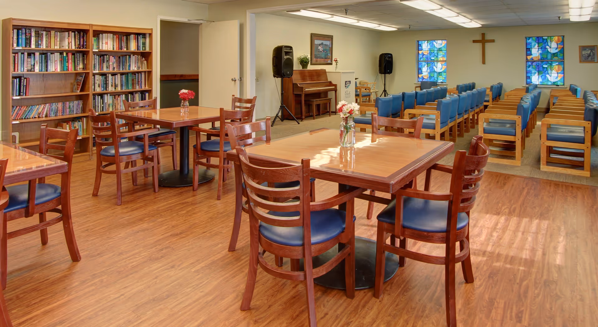 A spacious room with wooden tables and chairs arranged on a wood floor. There are bookshelves filled with books on the left side and a piano with speakers in the back. Rows of blue cushioned chairs face forward towards a wall with two stained glass windows and a wooden cross mounted between them.
