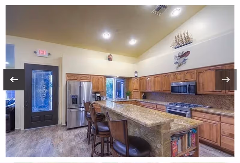 A spacious kitchen with wooden cabinets, a large granite island with bar stools, stainless steel refrigerator, stove, and microwave. The kitchen has high ceilings with recessed lighting and decorative items on top of the cabinets. There is a door with a decorative glass panel and an exit sign above it.