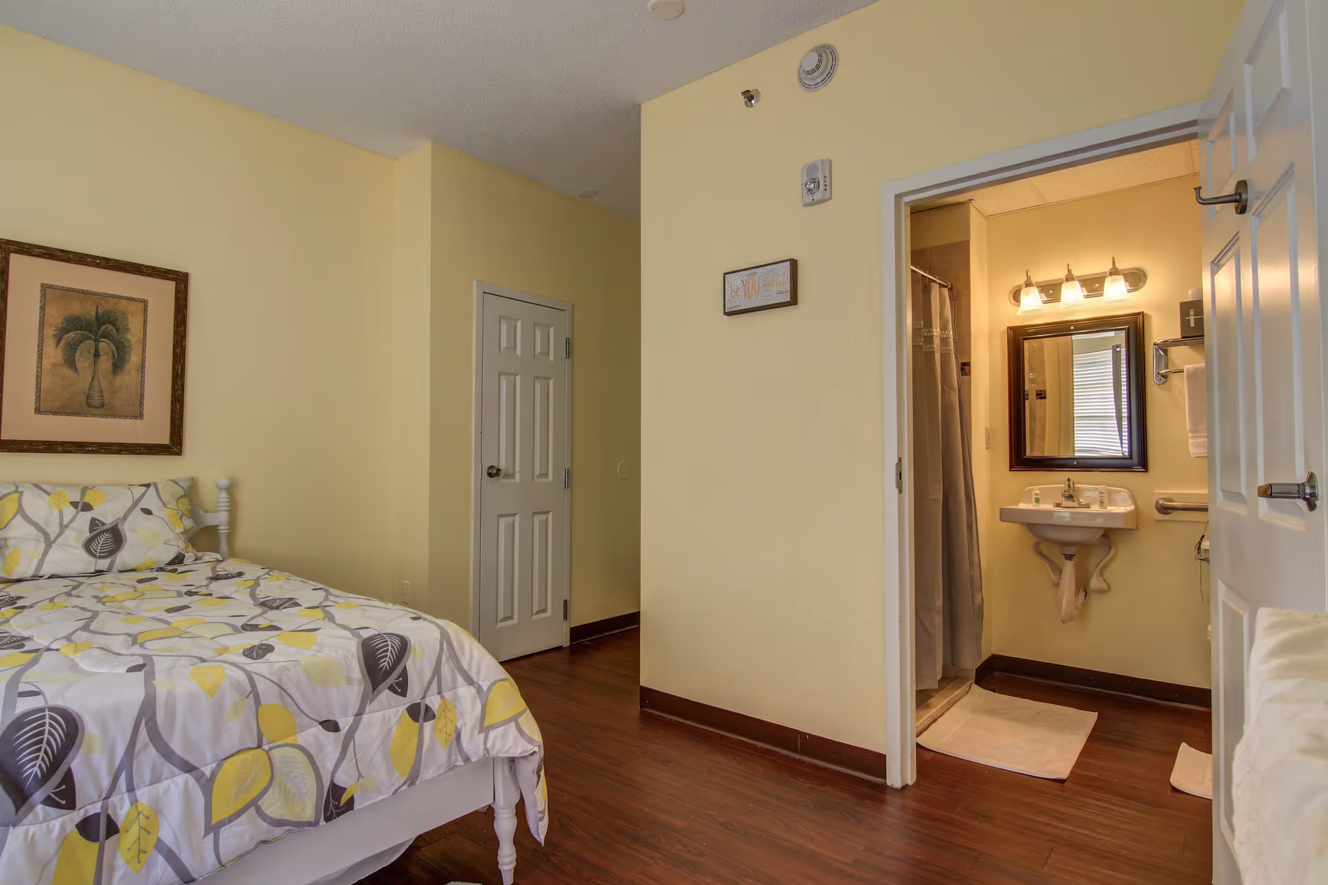Small bedroom with a patterned bed, framed artwork, and an open door revealing a bathroom with sink and mirror.