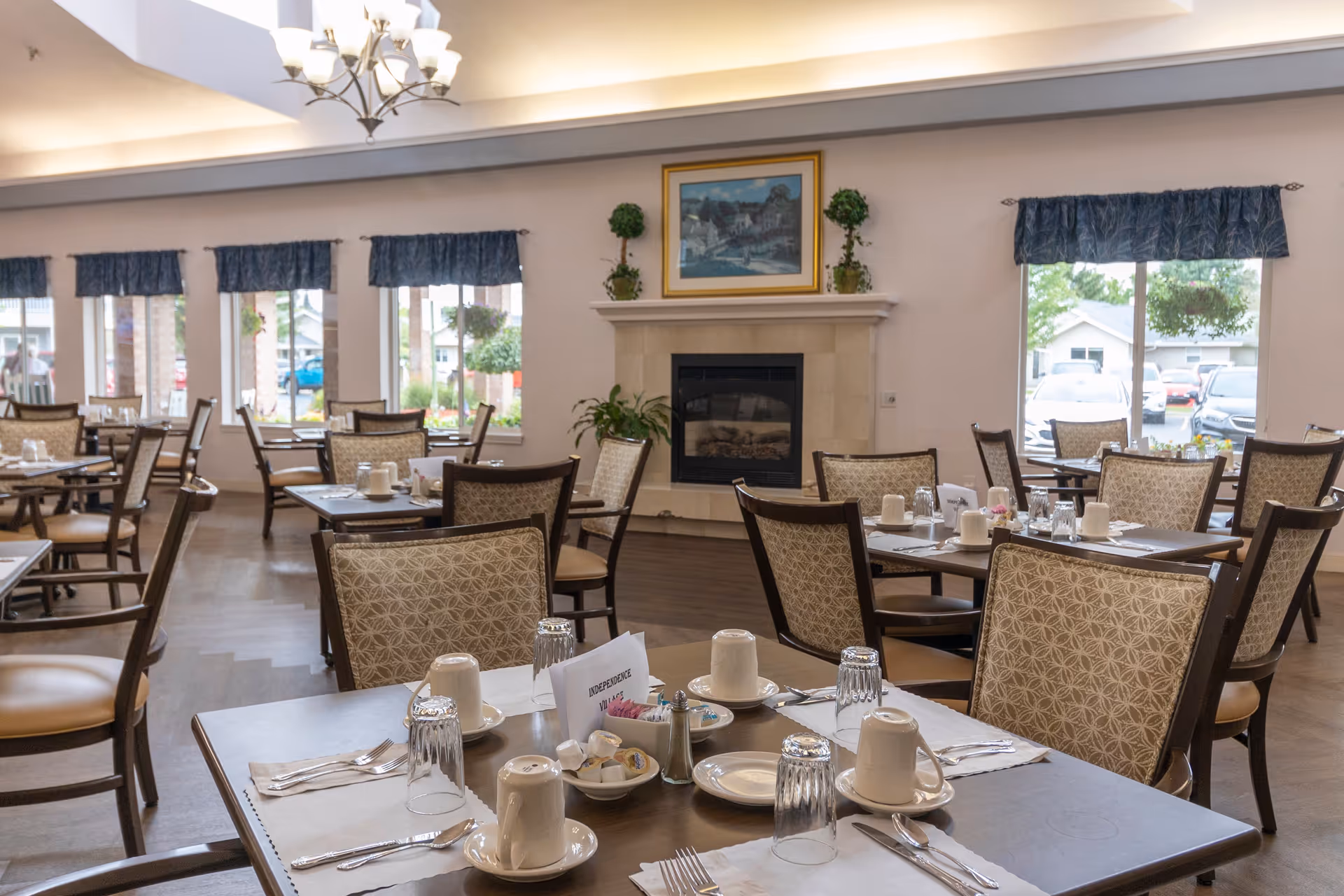 A dining room in a senior living facility with multiple tables set for meals. Each table has cups, glasses, plates, utensils, and condiments. The room features large windows with blue valances, a fireplace with a framed painting above it, and potted plants on the mantel. Outside the windows, parked cars and houses are visible.