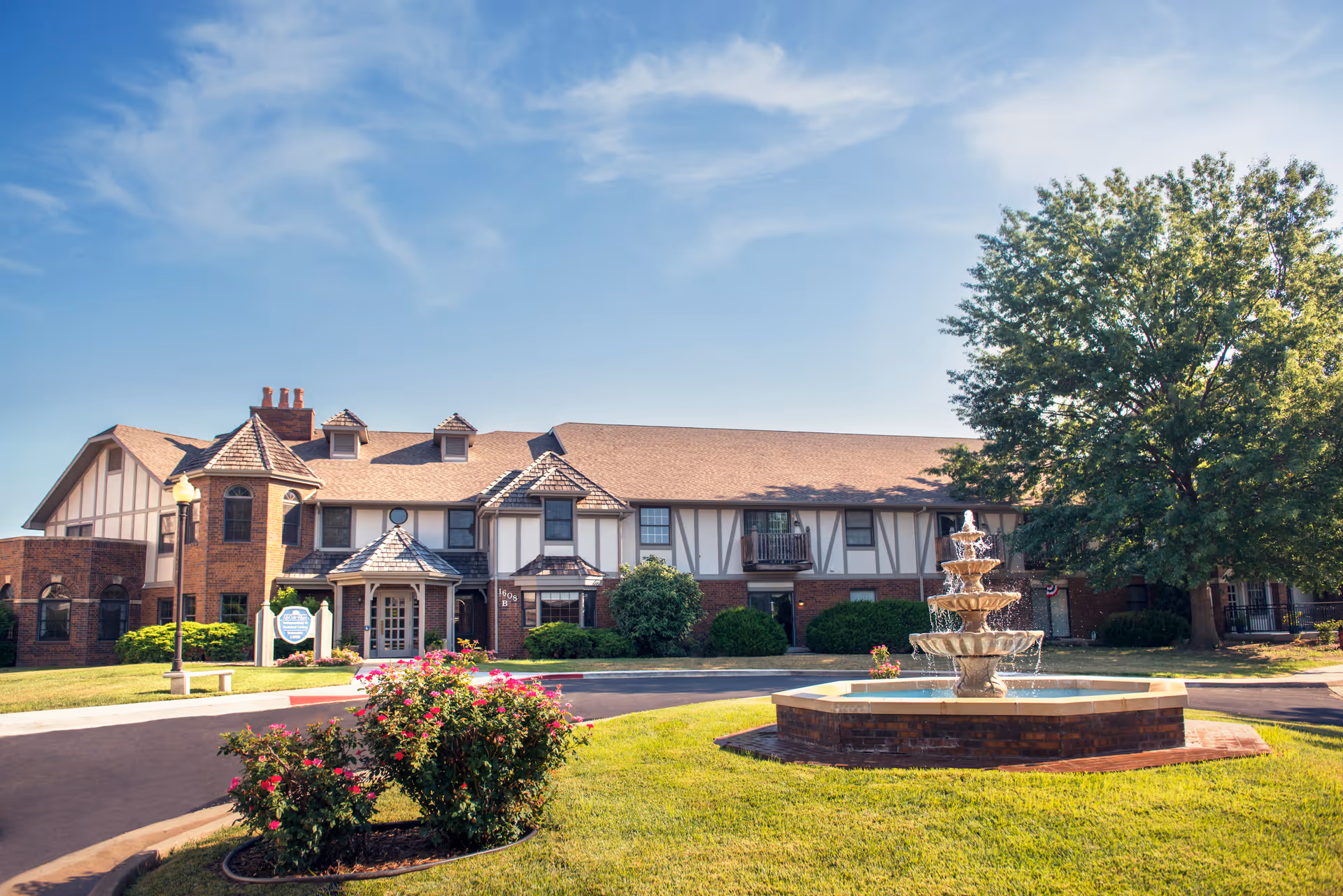 Two-story brick and Tudor-style building front with a tiered fountain and manicured lawn under a blue sky.