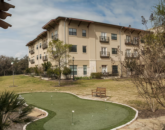 Three-story beige assisted living building beside a small putting green and a bench on a grassy lawn.