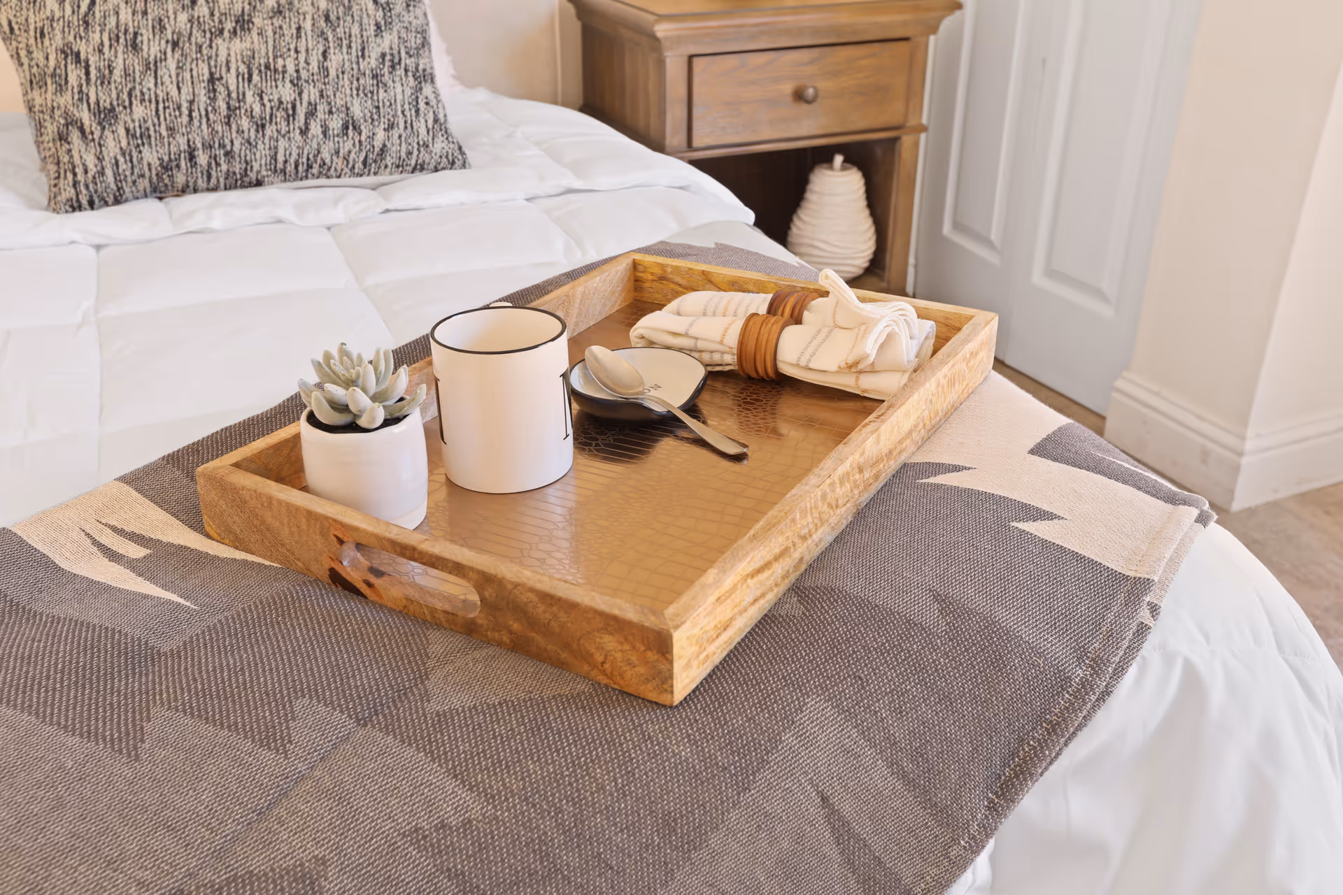 A wooden serving tray holding a small succulent, a mug, utensils and a napkin sits on a patterned throw at the foot of a neatly made bed.