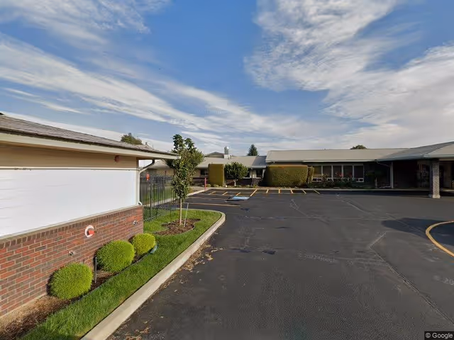 Parking lot and entrance area of a single-story assisted living building under a partly cloudy sky.