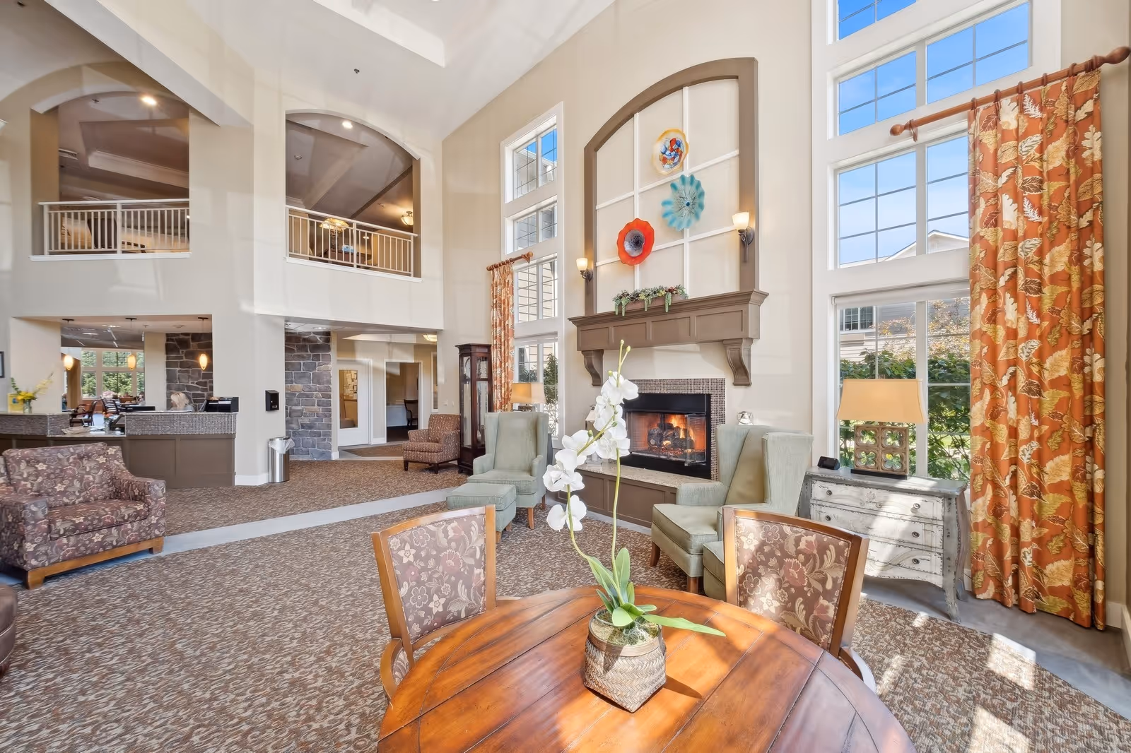 A spacious and well-lit living room area in a senior living facility with high ceilings and large windows. The room features a fireplace with decorative wall art above it, two green armchairs, a floral-patterned armchair, a wooden round table with a potted orchid, and a side table with a lamp. The room has patterned carpet and orange floral curtains on the windows.