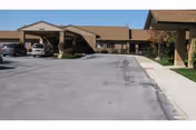 Exterior view of a single-story senior living facility with a paved driveway and parked vehicles in front. The building has a brown roof and brick walls, with a covered entrance area.