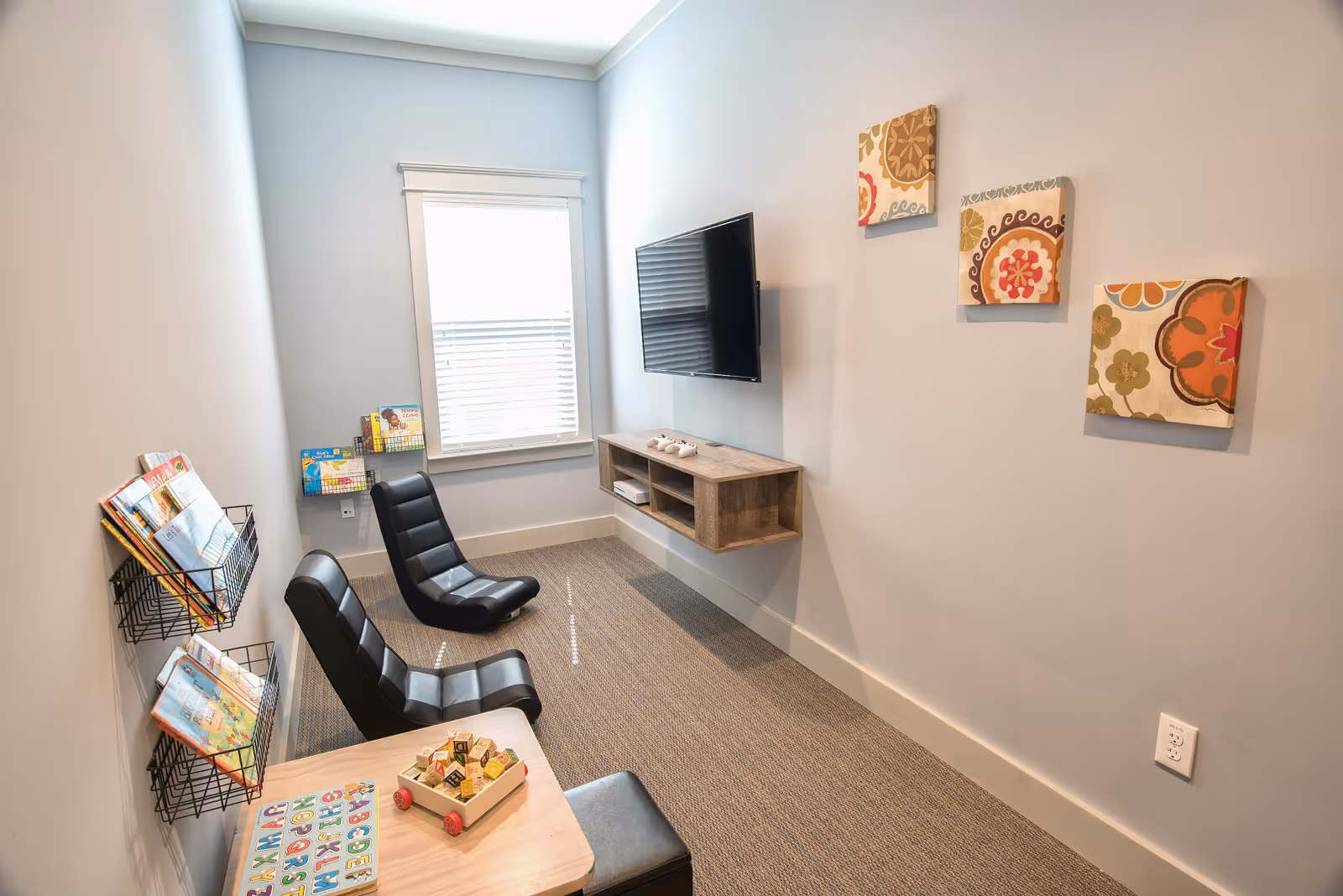 A small, cozy room with two black cushioned floor chairs facing a wall-mounted flat-screen TV. Below the TV is a floating wooden shelf with gaming controllers. On the left wall, there are wire racks holding children's books. A small wooden table with alphabet blocks and toys is in the foreground. The walls are light gray with three colorful floral-patterned square canvases hanging on the right wall. A window with blinds is on the far wall, letting in natural light.