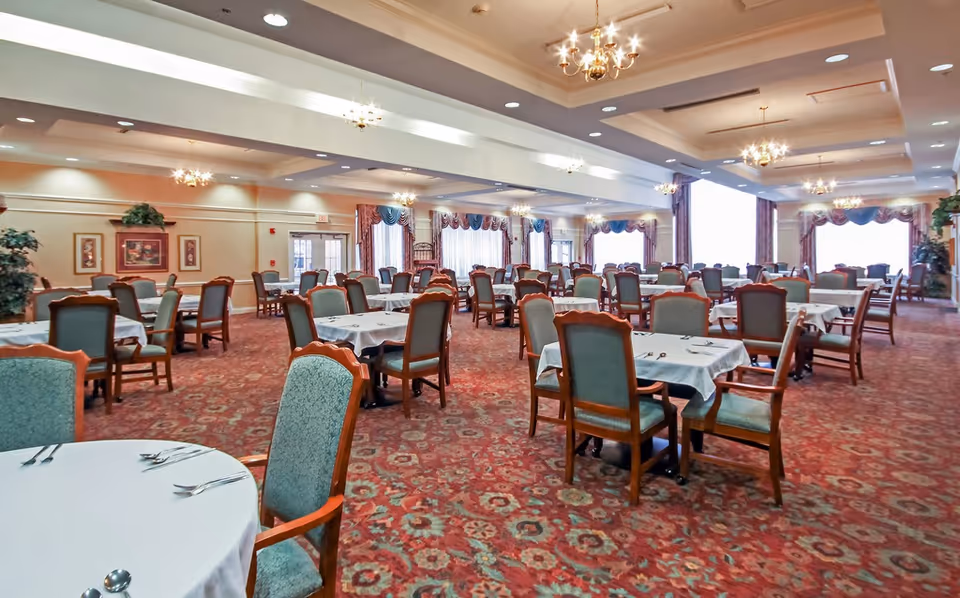 Large formal dining room with multiple round and rectangular tables covered in white tablecloths and upholstered chairs under chandeliers.