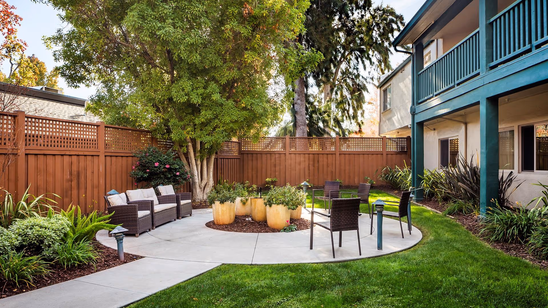 Outdoor garden area at Atria Sunnyvale featuring a circular concrete patio with several potted plants in the center, surrounded by wicker chairs and cushioned seating. The space is enclosed by a wooden fence with greenery and trees providing shade and a serene atmosphere.