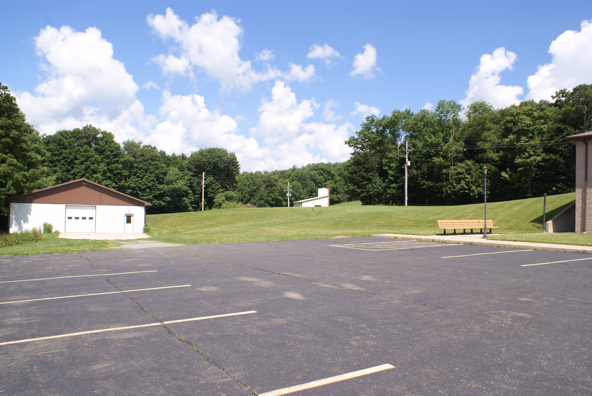 Empty parking lot with a grassy hill, a small garage building and a bench in front of a tree line under a blue sky.