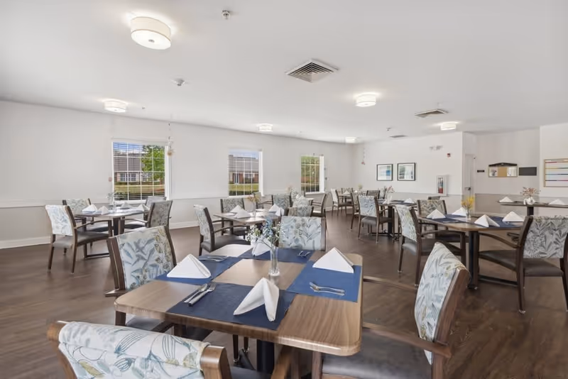 A bright and spacious dining room with multiple wooden tables set with blue placemats, white folded napkins, silverware, and small floral centerpieces. The room has large windows with white blinds, light-colored walls, and wooden flooring. Comfortable chairs with floral patterned upholstery surround each table.