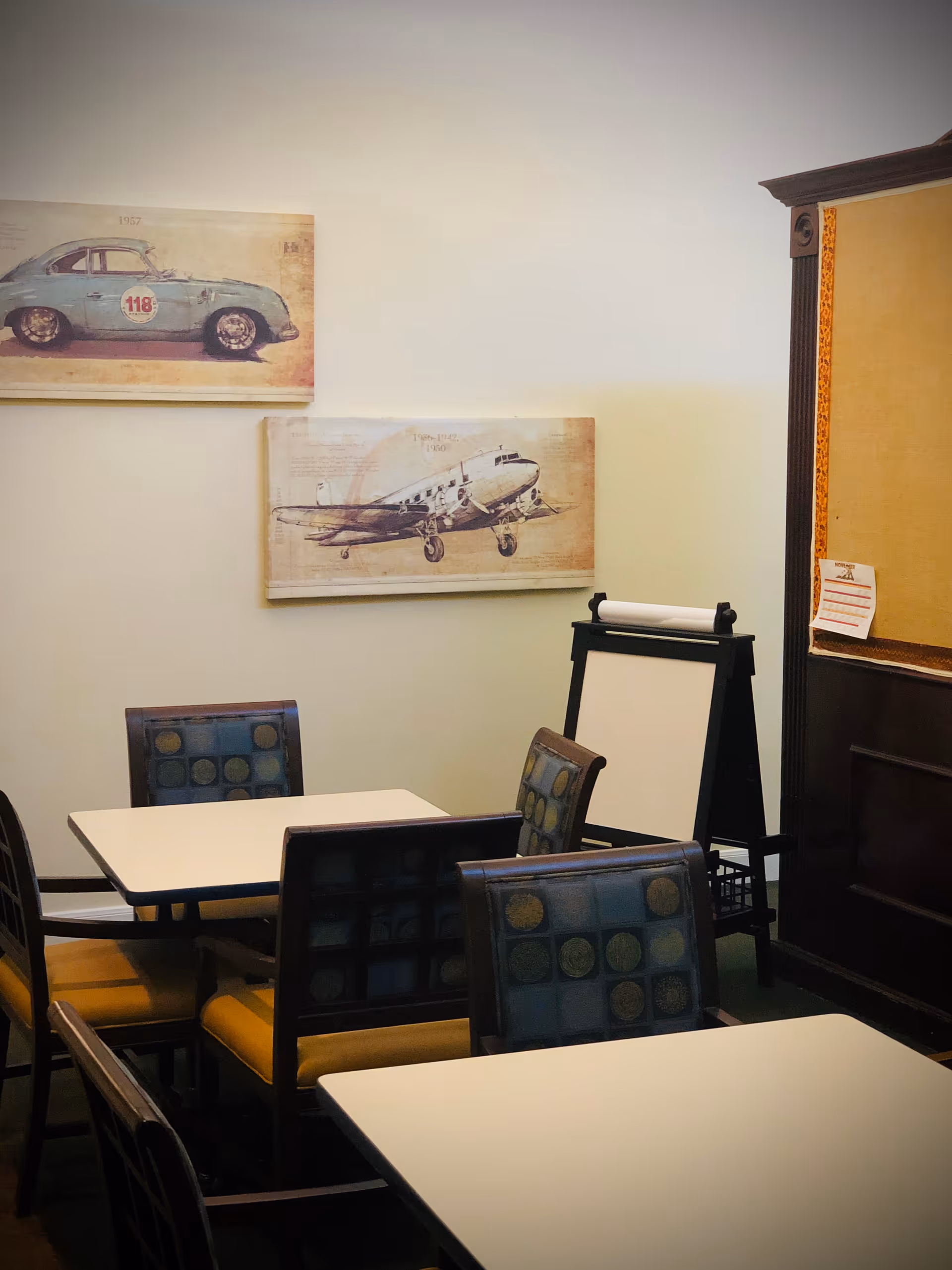 A small room with several square tables and chairs with patterned backs and yellow seats. On the wall are two vintage-style paintings, one of a blue race car and the other of an airplane. There is also a blank easel and a bulletin board with a small paper pinned to it.