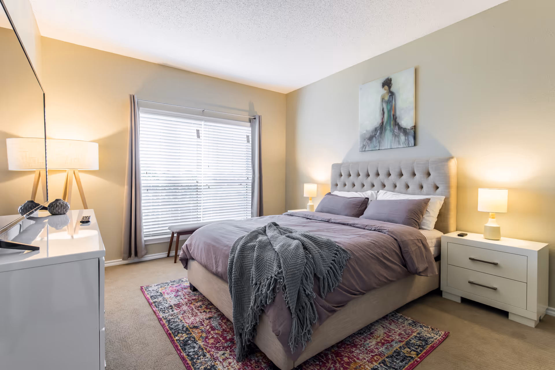 Well-lit bedroom with a tufted bed, bedside tables and lamps, dresser, window with blinds, and a colorful rug.