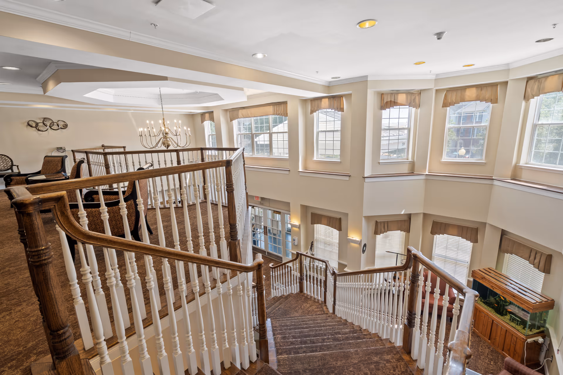 Interior view of a senior living facility showing a wooden staircase with white balusters leading down to a lower level. The area is well-lit with multiple windows featuring beige valances. There is a chandelier hanging from the ceiling, several chairs arranged on the upper level, and a fish tank on the lower level near the windows.