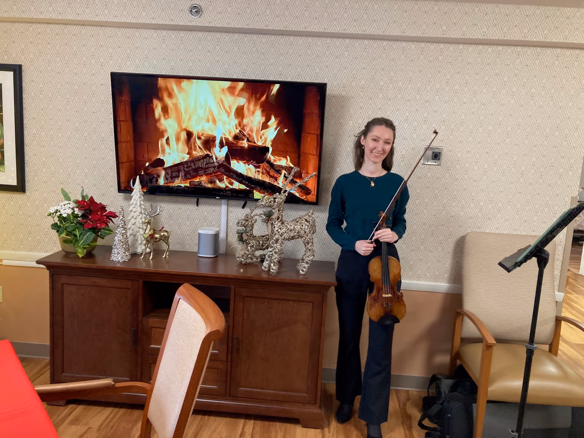 A person standing in a lounge holding a violin beside a TV displaying a fireplace and holiday decorations on a wooden sideboard.
