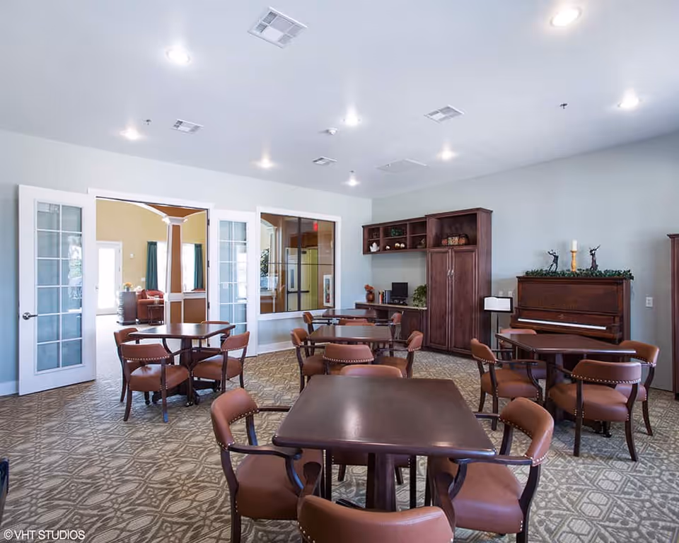 A well-lit common room with multiple wooden tables and brown cushioned chairs arranged neatly. The room features a patterned carpet, light-colored walls, and a piano against one wall decorated with greenery and candles. There are glass-paneled double doors leading to another room with more seating and windows with curtains.