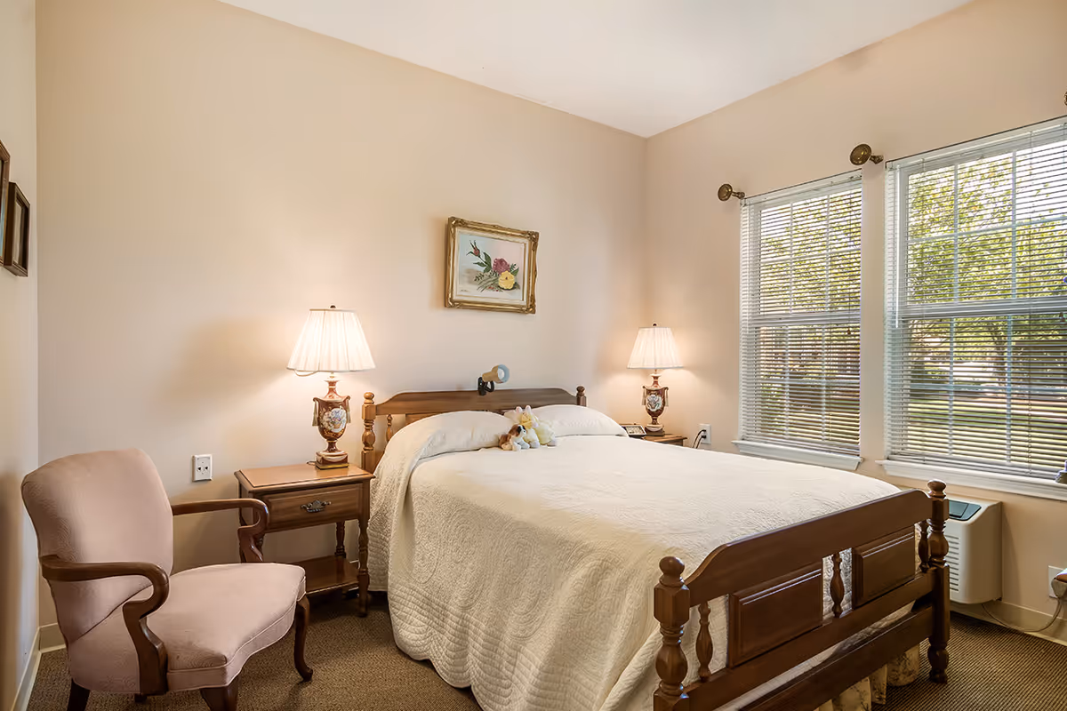 A cozy bedroom with a wooden bed frame covered with a white quilt. Two bedside tables each hold a decorative lamp. A framed floral painting hangs above the bed. There is a pink upholstered armchair to the left and two large windows with blinds on the right letting in natural light.