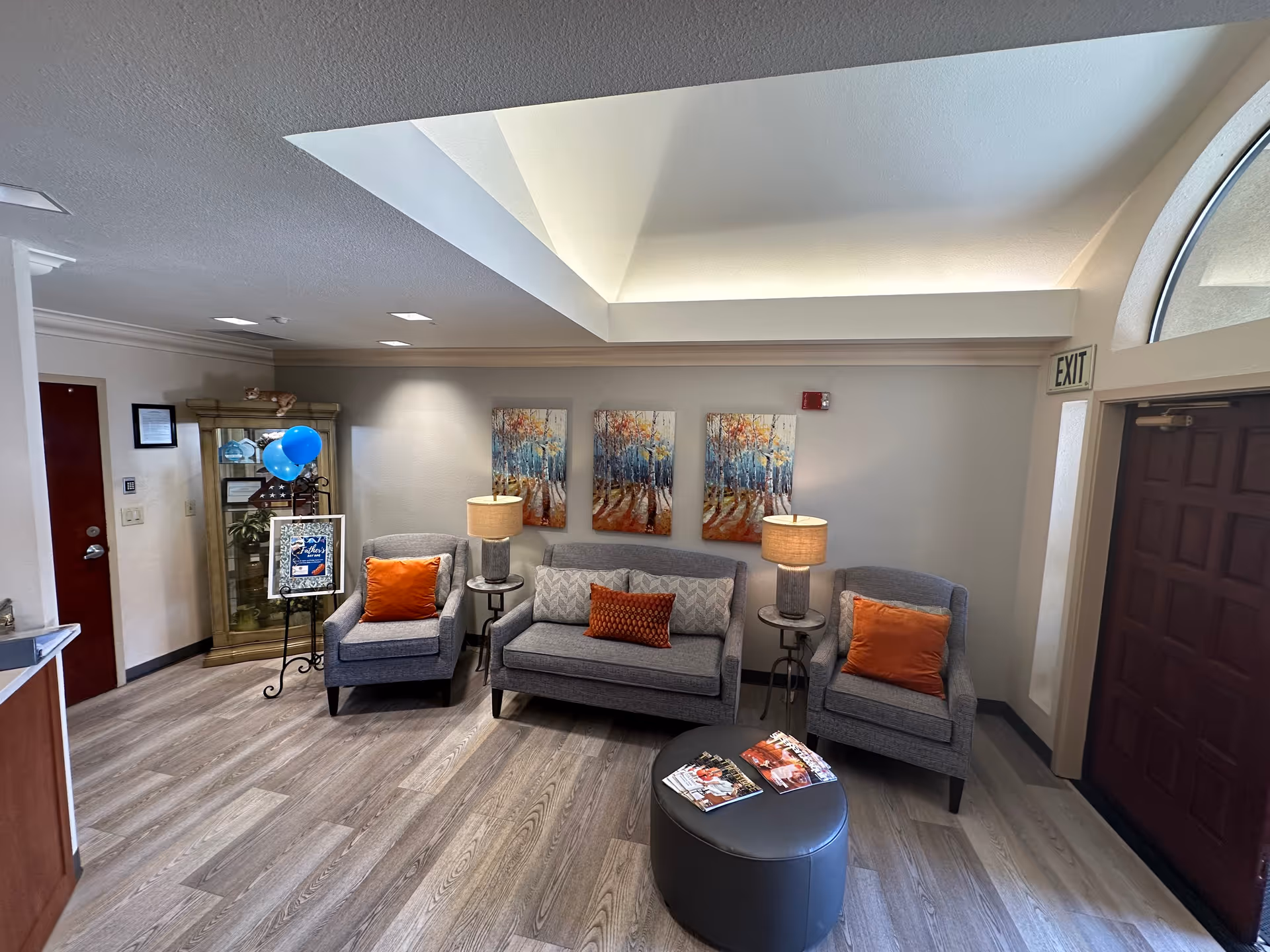 A welcoming lobby area featuring gray seating with orange cushions, a round coffee table, and decorative artwork on the walls. A blue balloon and a sign are also visible, along with an exit door.