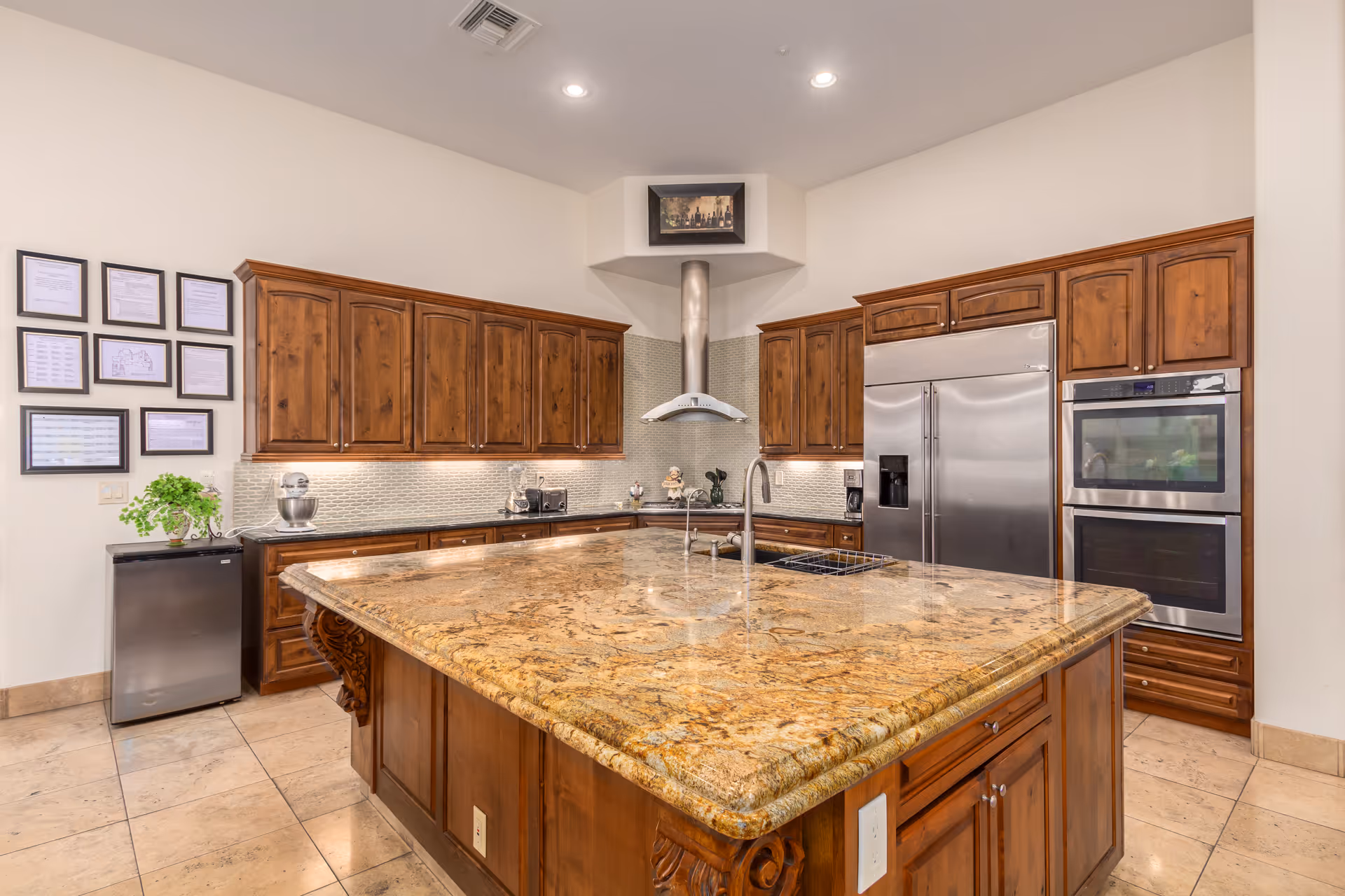 A spacious kitchen with a large granite island countertop in the center, surrounded by wooden cabinets. The kitchen features stainless steel appliances including a double oven and a refrigerator. There is a modern range hood above the stove area and framed certificates or documents on the wall to the left. The floor is tiled and the lighting is bright with recessed ceiling lights.