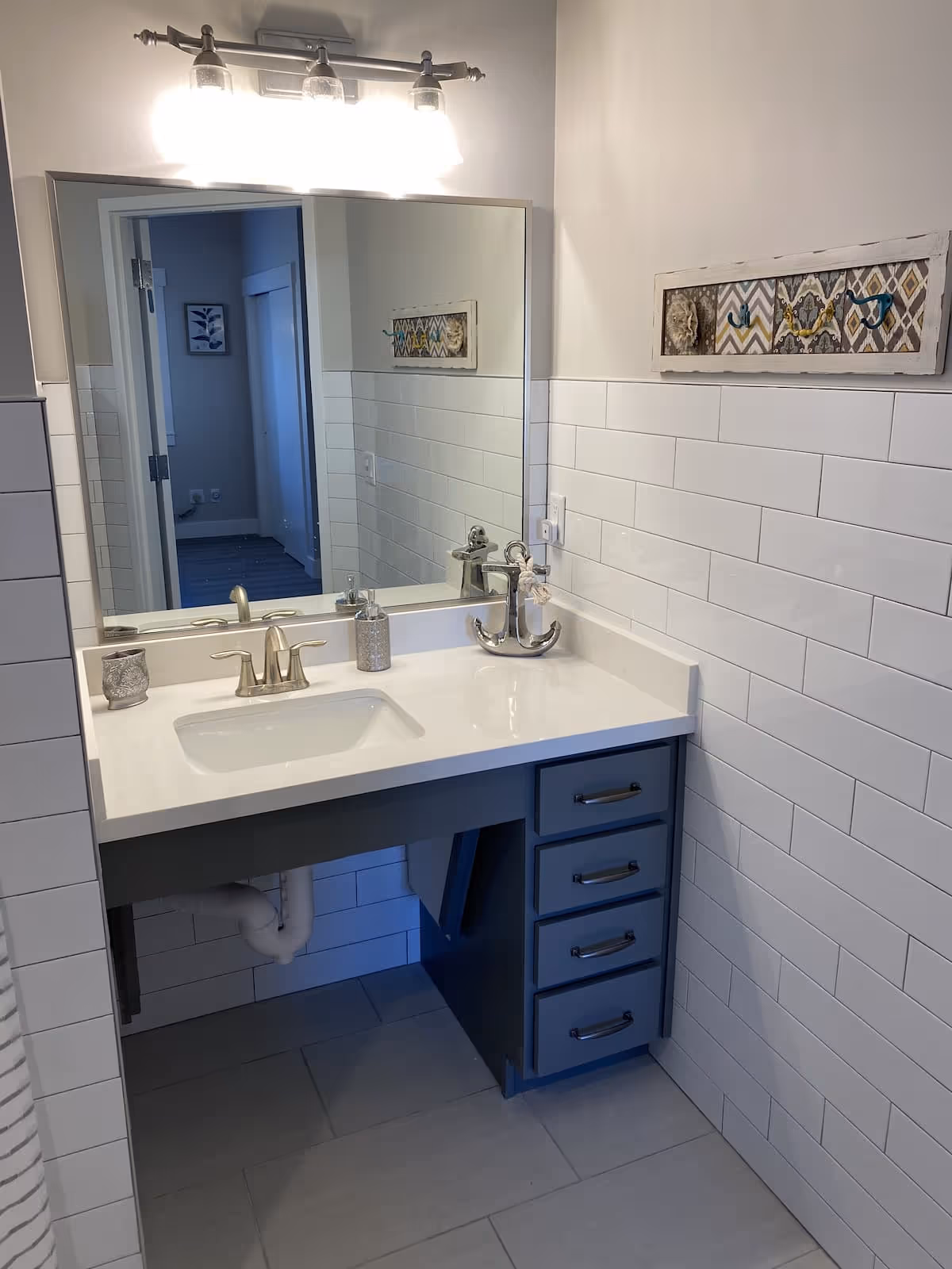 A bathroom vanity with a white countertop, a built-in sink, and a silver faucet. Above the vanity is a large rectangular mirror with a three-light fixture mounted on the wall. The walls are partially tiled with white subway tiles, and there is a decorative wall hanging with hooks on the right side. The vanity has four drawers on the right side and an open space underneath the sink. The floor is tiled in light gray.