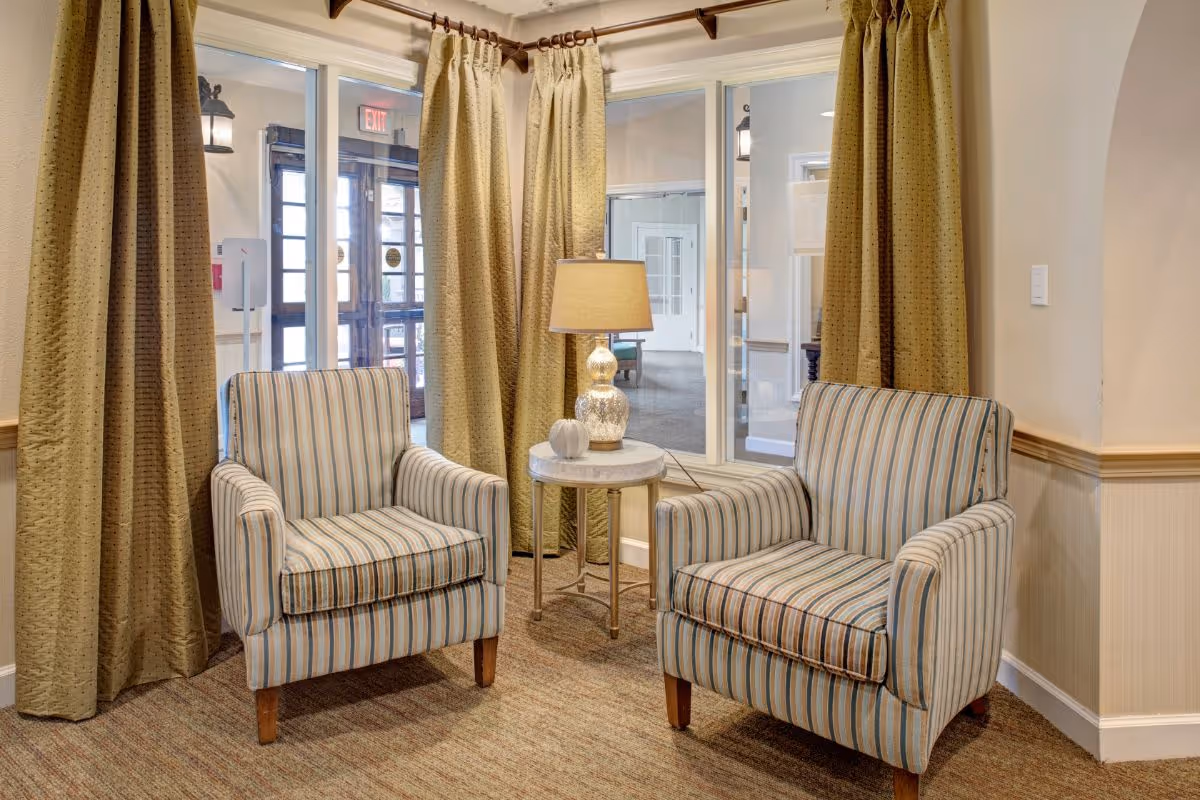 Two striped armchairs with wooden legs are positioned on either side of a small round table with a lamp and a decorative white pumpkin on it. Behind the chairs are large windows with beige curtains, and through the windows, an exit door and part of a hallway are visible.