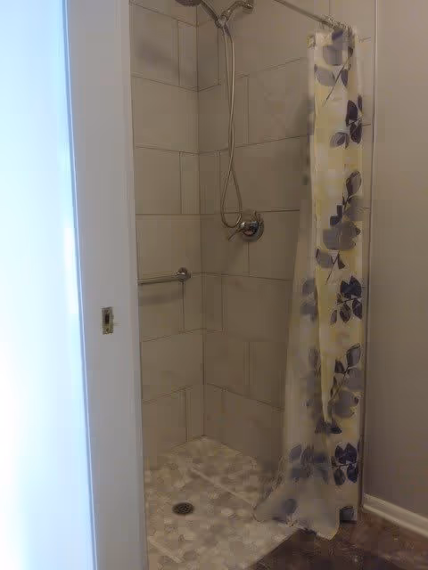 A tiled shower area with a silver showerhead and a floral-patterned shower curtain partially drawn to the side. The shower floor has hexagonal tiles and there is a metal grab bar on the wall.
