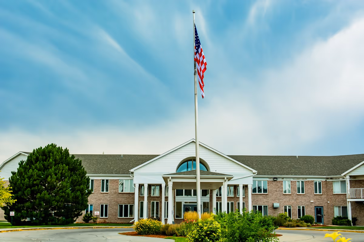 Front exterior view of a two-story brick building with white trim and a covered entrance supported by columns. An American flag is flying on a flagpole in front of the building, surrounded by landscaped bushes and flowers under a partly cloudy sky.