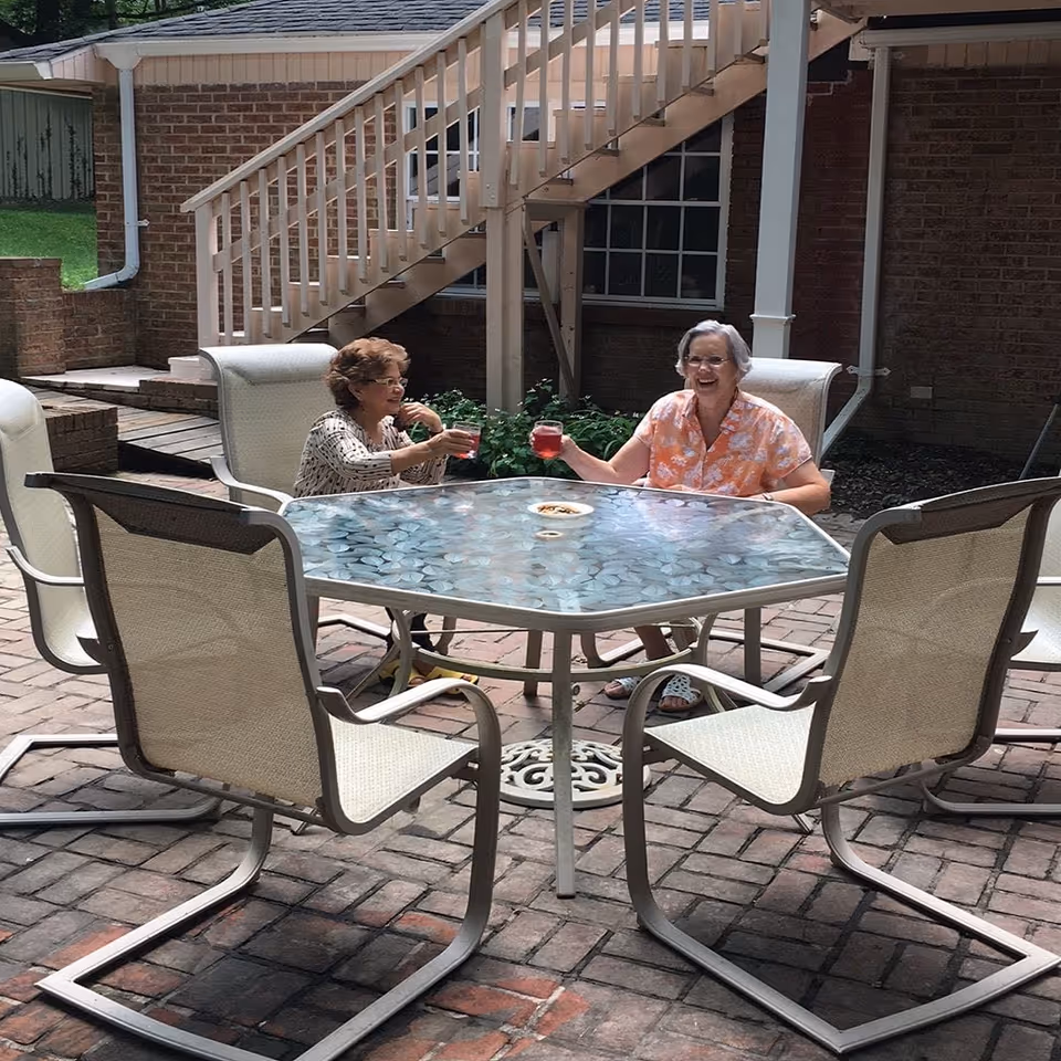 Two elderly women sitting at a glass-top outdoor table on a brick patio, smiling and clinking glasses with a brick building and staircase in the background.