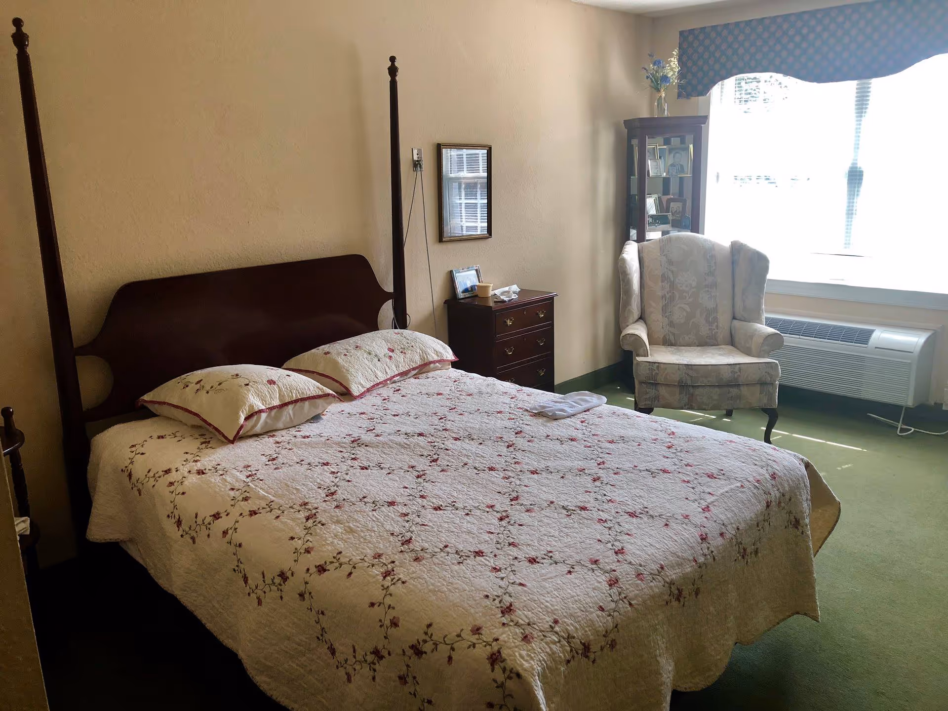 A furnished bedroom with a four-poster bed covered in a floral quilt, a nightstand and dresser, an upholstered armchair by a bright window, and a wall air conditioner.