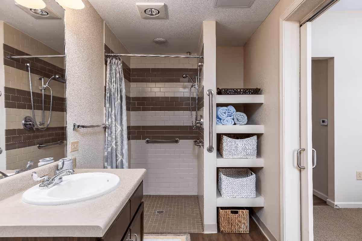 A bathroom with a sink and countertop on the left, a walk-in shower with a curtain and grab bars in the center, and built-in shelves on the right holding rolled towels and baskets. The bathroom has beige and brown tiled walls and a sliding door leading to another room.