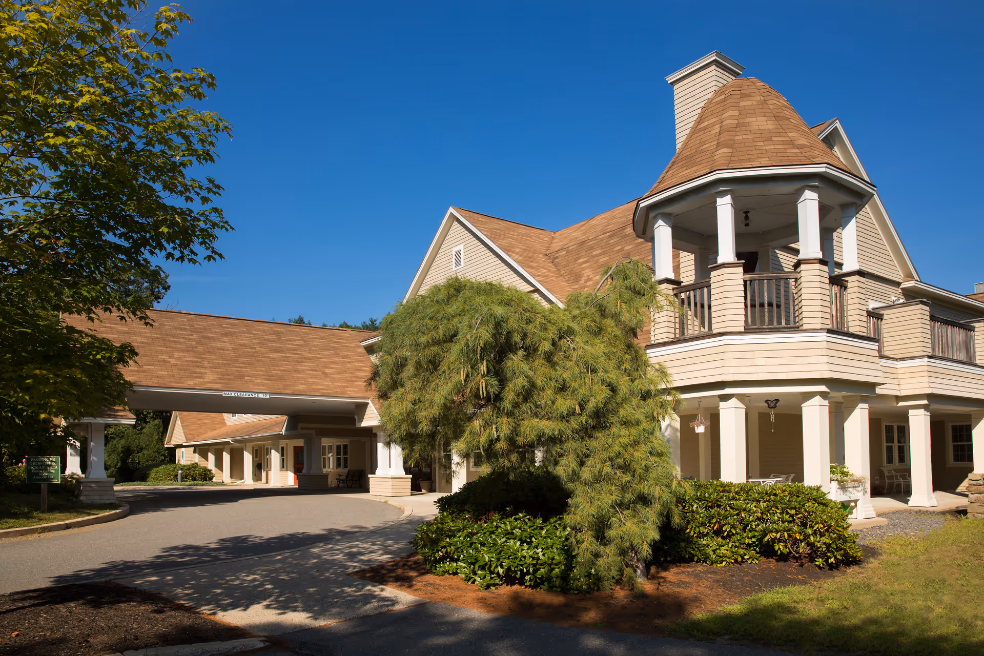 Exterior view of a senior living facility building with beige siding and a brown shingled roof under a clear blue sky. The building features a covered driveway entrance, a turret with a balcony, and surrounding greenery including bushes and trees.