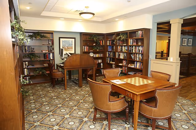A cozy library or reading room with wooden bookshelves filled with books and decorative plants. There is a wooden desk with a computer and chair, and a square wooden table with a built-in chessboard surrounded by four leather chairs. The room has patterned carpet flooring and warm lighting.