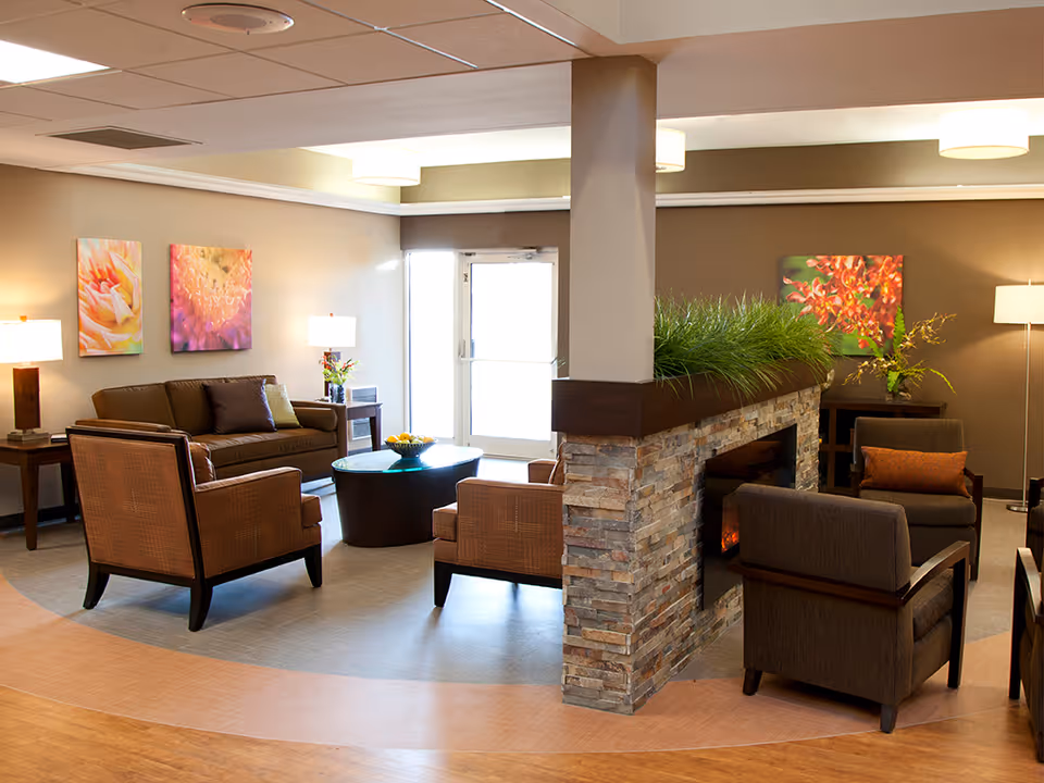 A cozy living room area in Greenbriar Healthcare Center featuring a stone fireplace with green plants on top, surrounded by several cushioned chairs and a sofa. The room is decorated with colorful floral paintings on the walls and illuminated by ceiling lights and floor lamps. A round coffee table with a bowl of fruit is placed in the center.