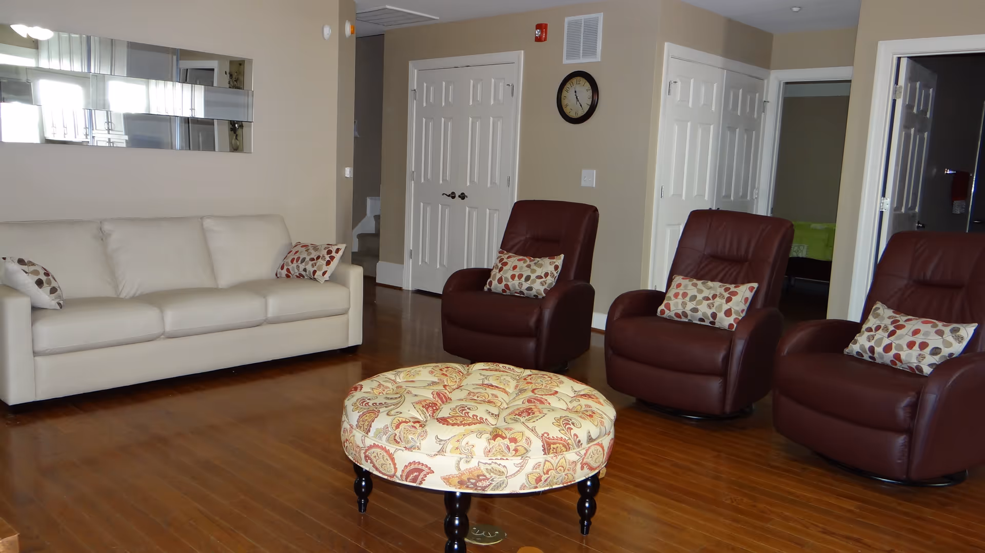 A living room with a beige sofa and three brown leather armchairs, each with a decorative pillow. In the center is a round, patterned ottoman. The room has wooden flooring, beige walls, a wall clock, and multiple white doors.