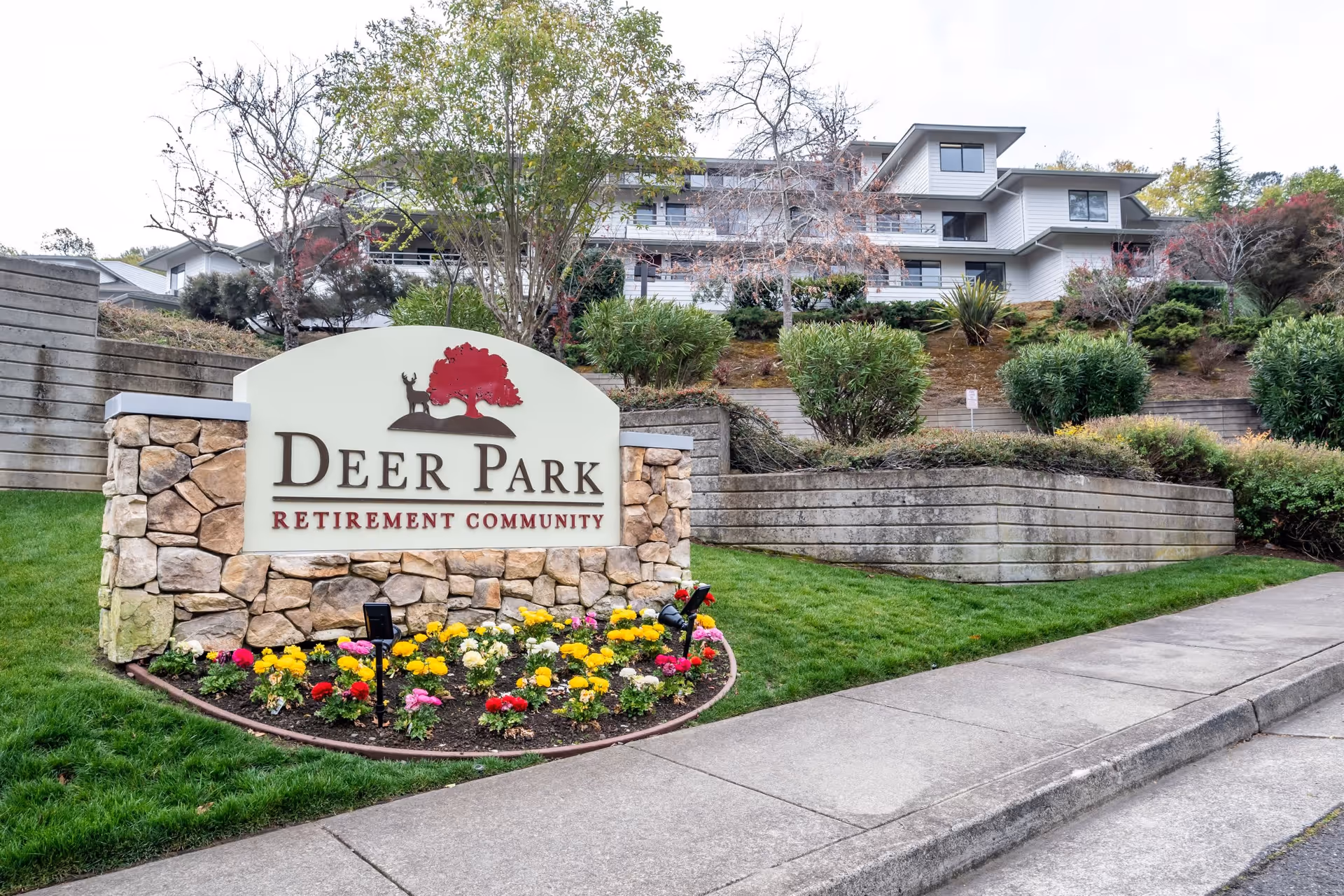 Entrance sign for Deer Park Retirement Community set in a stone structure with colorful flowers planted at its base. Behind the sign, there is a multi-story building surrounded by landscaped greenery and trees.