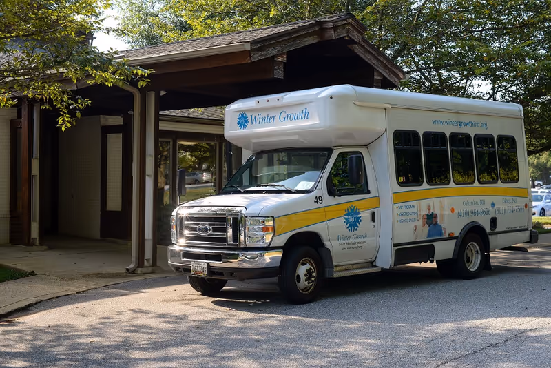 A white shuttle bus with Winter Growth branding parked outside a building entrance with a wooden overhang and surrounded by trees.