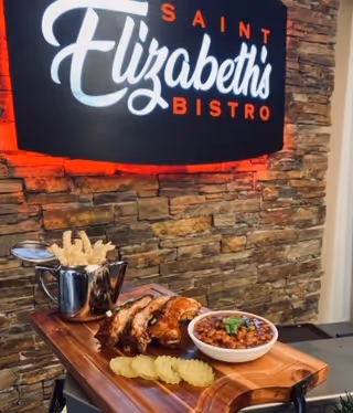 A wooden serving board with a meal consisting of ribs, a bowl of baked beans garnished with green onions, pickle slices, and a metal cup holding crispy fried snacks. The background features a stone wall with a sign that reads 'Saint Elizabeth's Bistro' illuminated with red lighting.