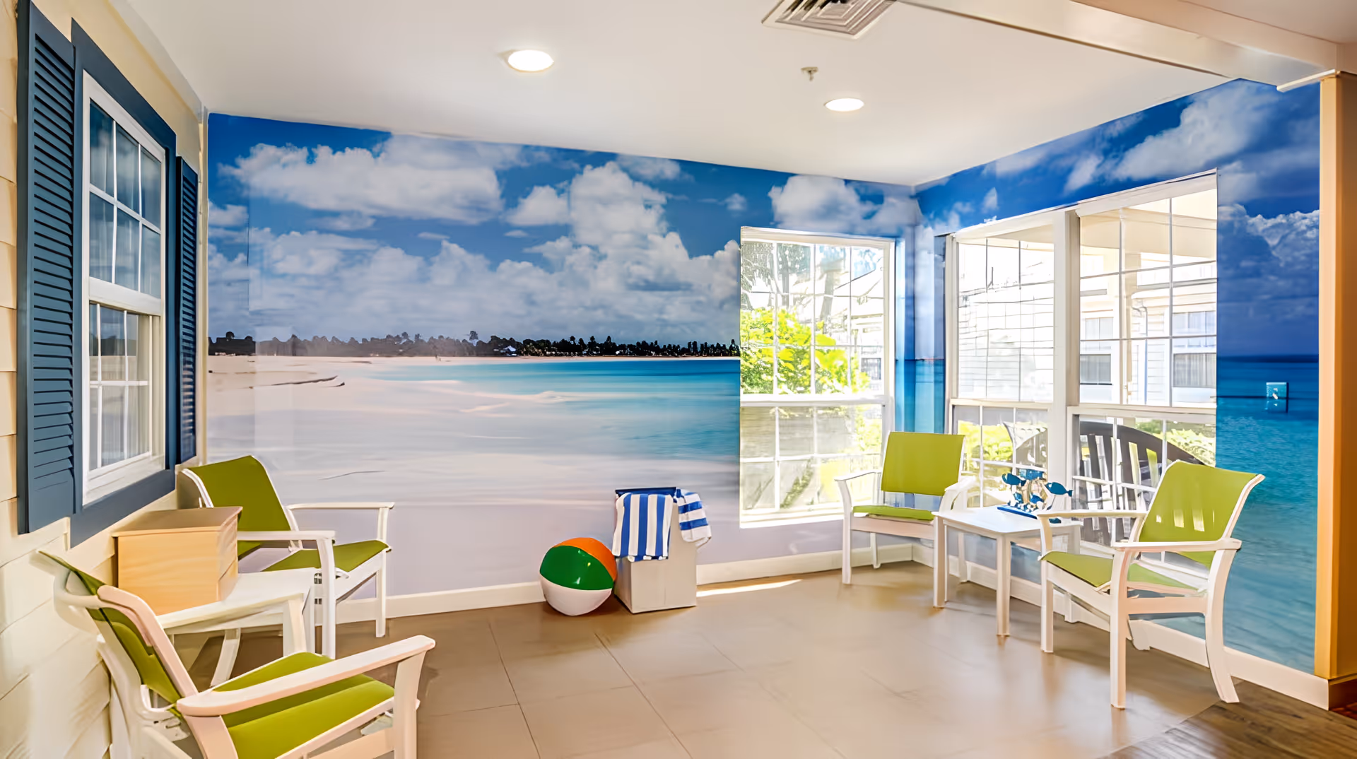 Sunlit common room with green-and-white chairs around small tables, a large beach-themed mural on the walls, and a beach ball on the floor.