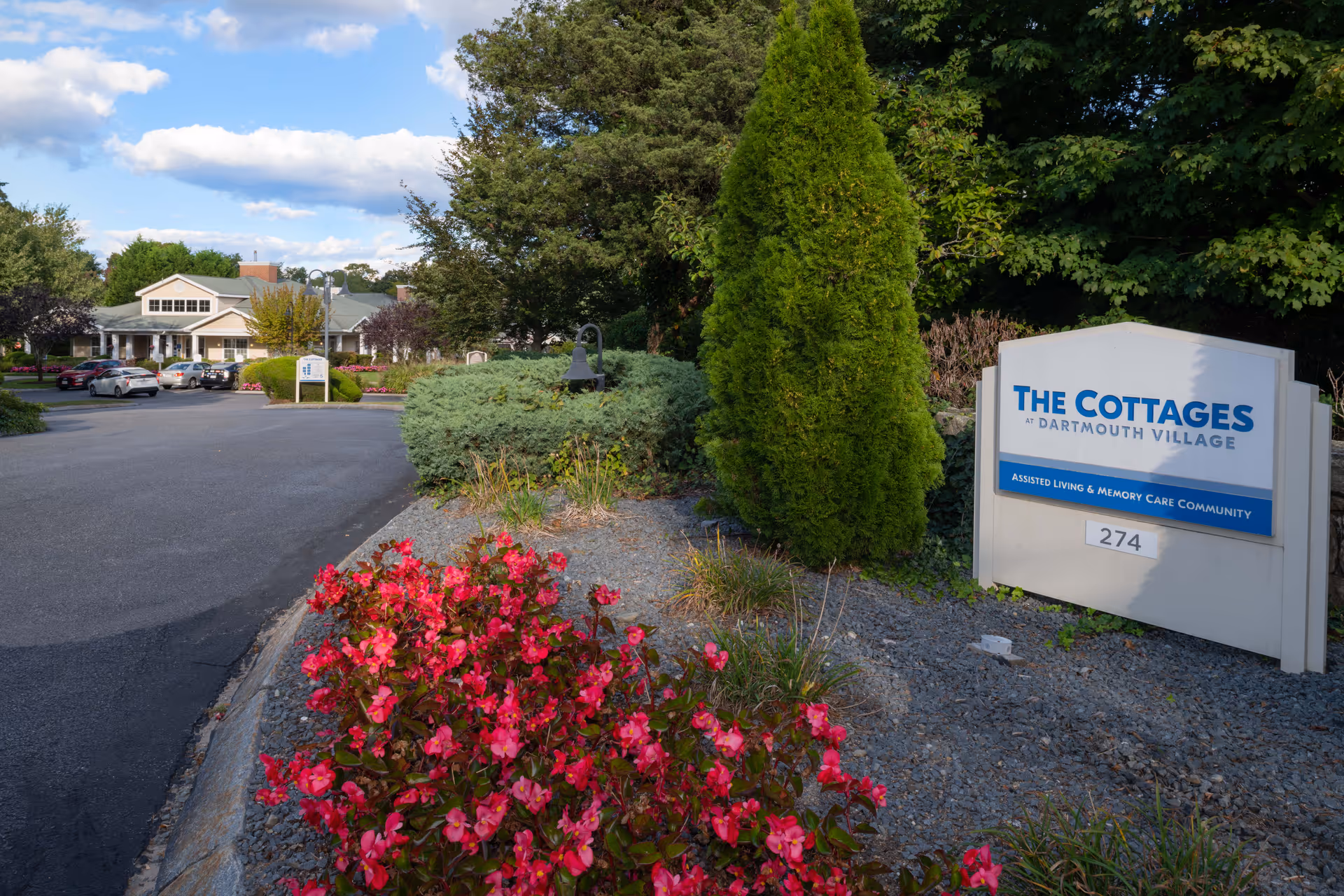 Entrance to The Cottages at Dartmouth Village assisted living and memory care community, featuring a sign with the facility name and address number 274, surrounded by landscaping with red flowers, bushes, and trees. In the background, there is a paved driveway leading to a building with parked cars and more greenery under a partly cloudy sky.