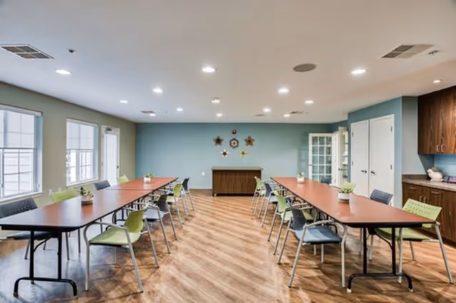 A spacious, well-lit room with two long tables arranged parallel to each other, surrounded by green and gray chairs. The room has large windows on one side, light wood flooring, and a blue accent wall with decorative wall hangings. There is a wooden cabinet against the far wall and built-in cabinets on the right side.