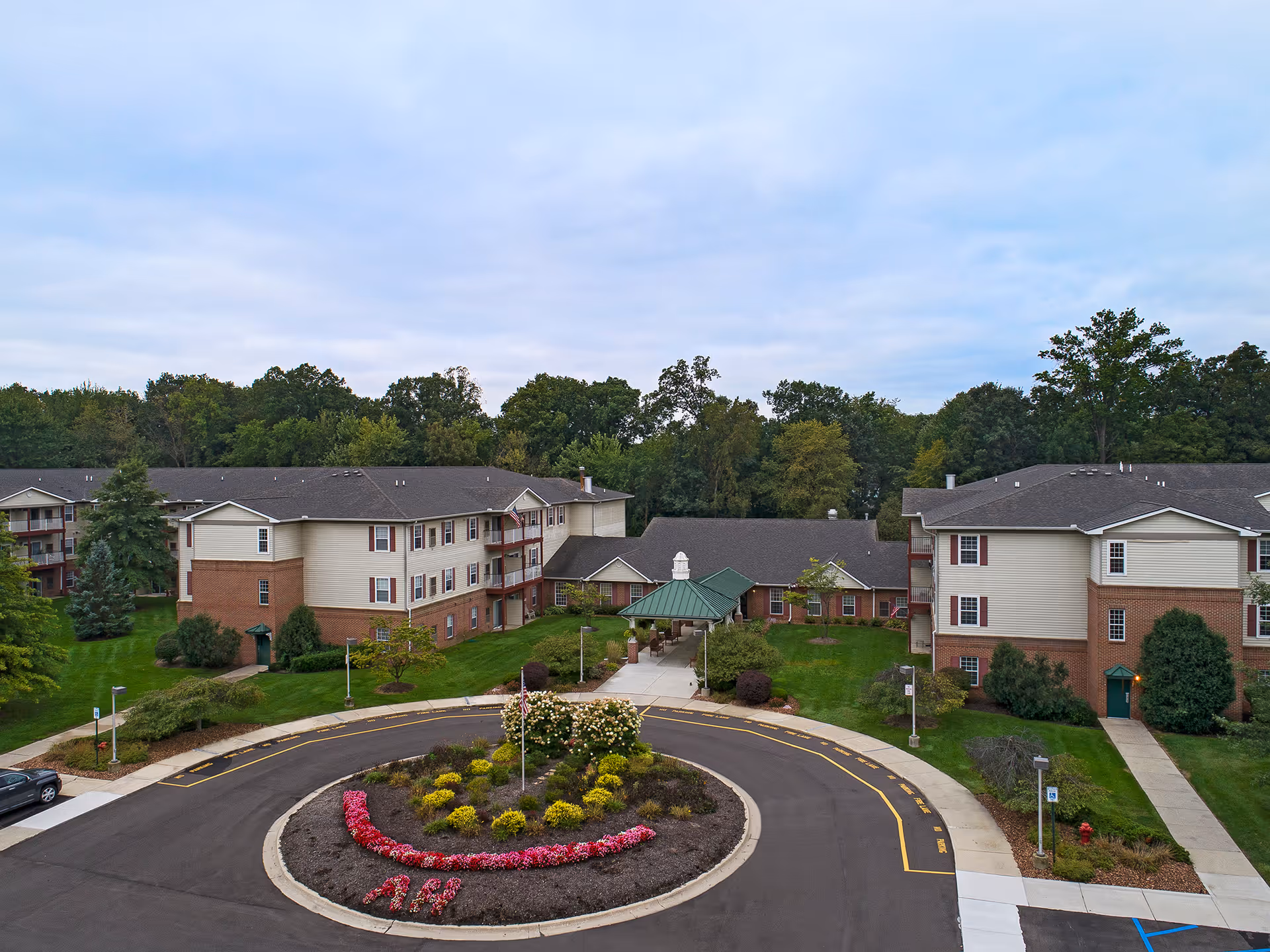 Front view of a senior living complex with a circular driveway, landscaped flower bed, and connected brick-and-siding buildings set against trees.