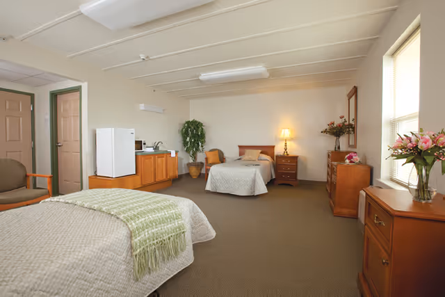 A spacious senior living bedroom with two single beds, each with beige bedspreads and one with a green throw blanket. The room features wooden furniture including dressers, a nightstand with a lamp, a chair, and a small kitchenette area with a mini fridge and microwave. There are flowers in vases on the dressers and a potted plant near the kitchenette. The room has beige walls, carpeted floor, and a window letting in natural light.
