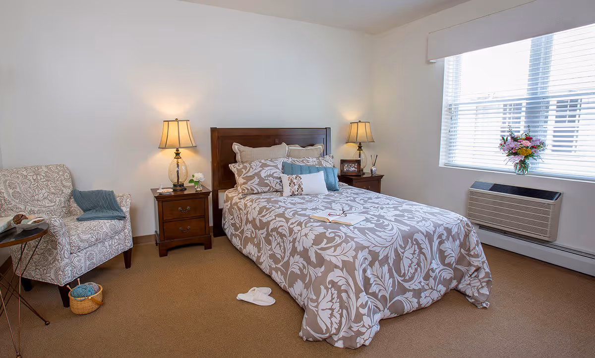 Neatly decorated bedroom with a bed centered between two nightstands and lamps, a patterned armchair, and a window with an air conditioner and flowers.