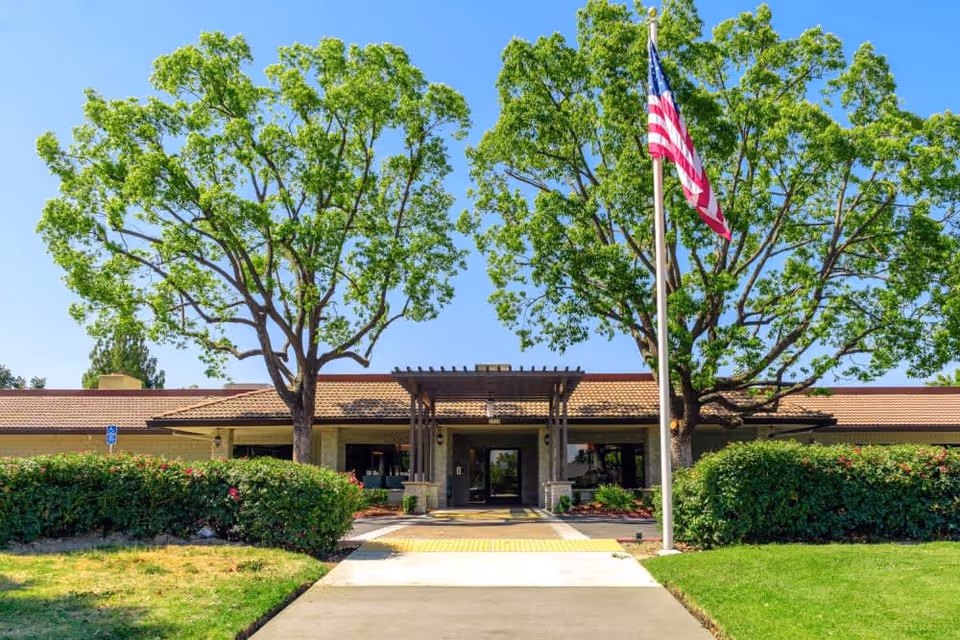Front exterior view of Park Visalia Assisted Living & Memory Care building with a pathway leading to the entrance, flanked by green bushes and two large trees. An American flag is displayed on a flagpole to the right of the entrance under a clear blue sky.